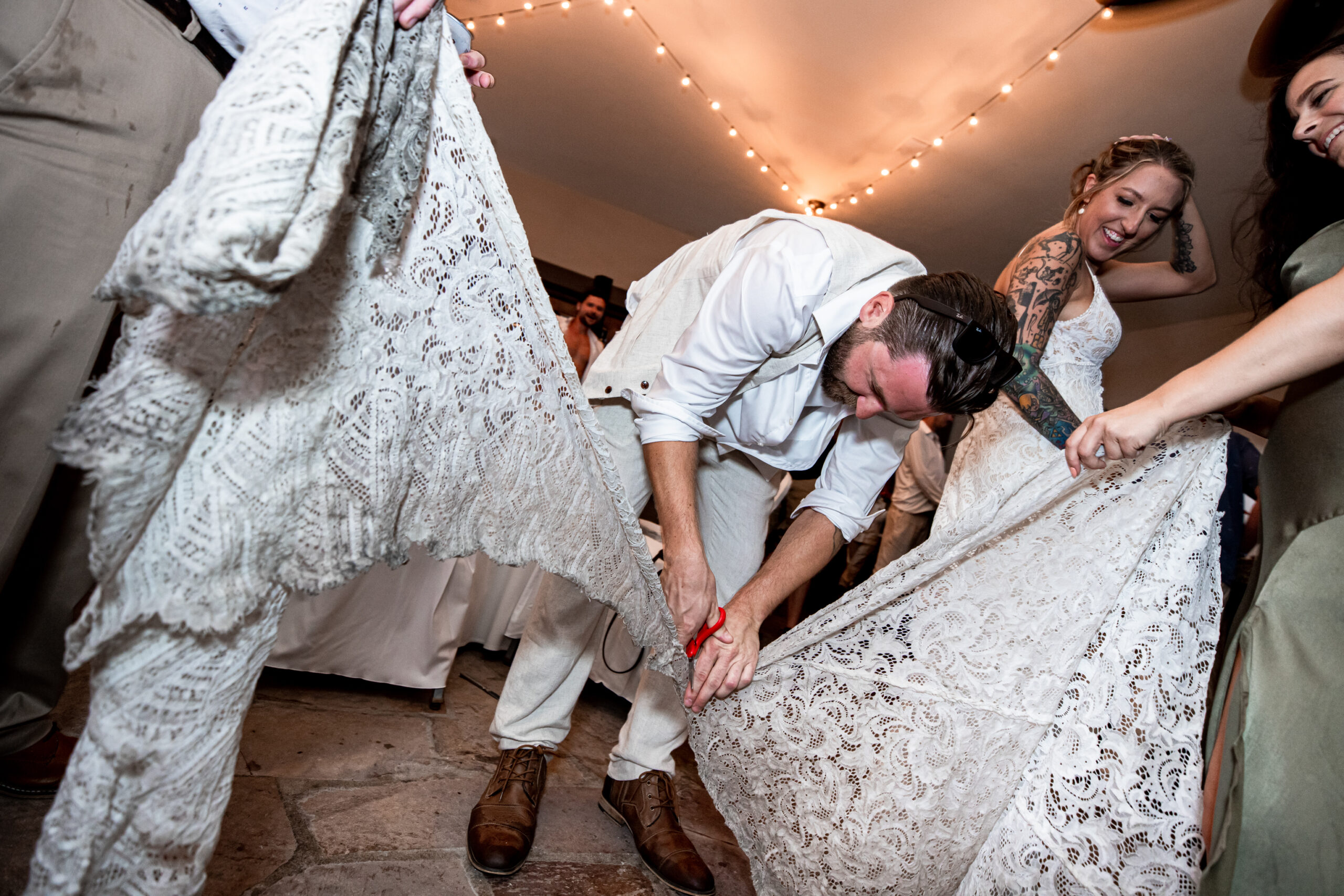 A bride looks down at her brother as he is cutting off the train of her dirty wedding dress with scissors.