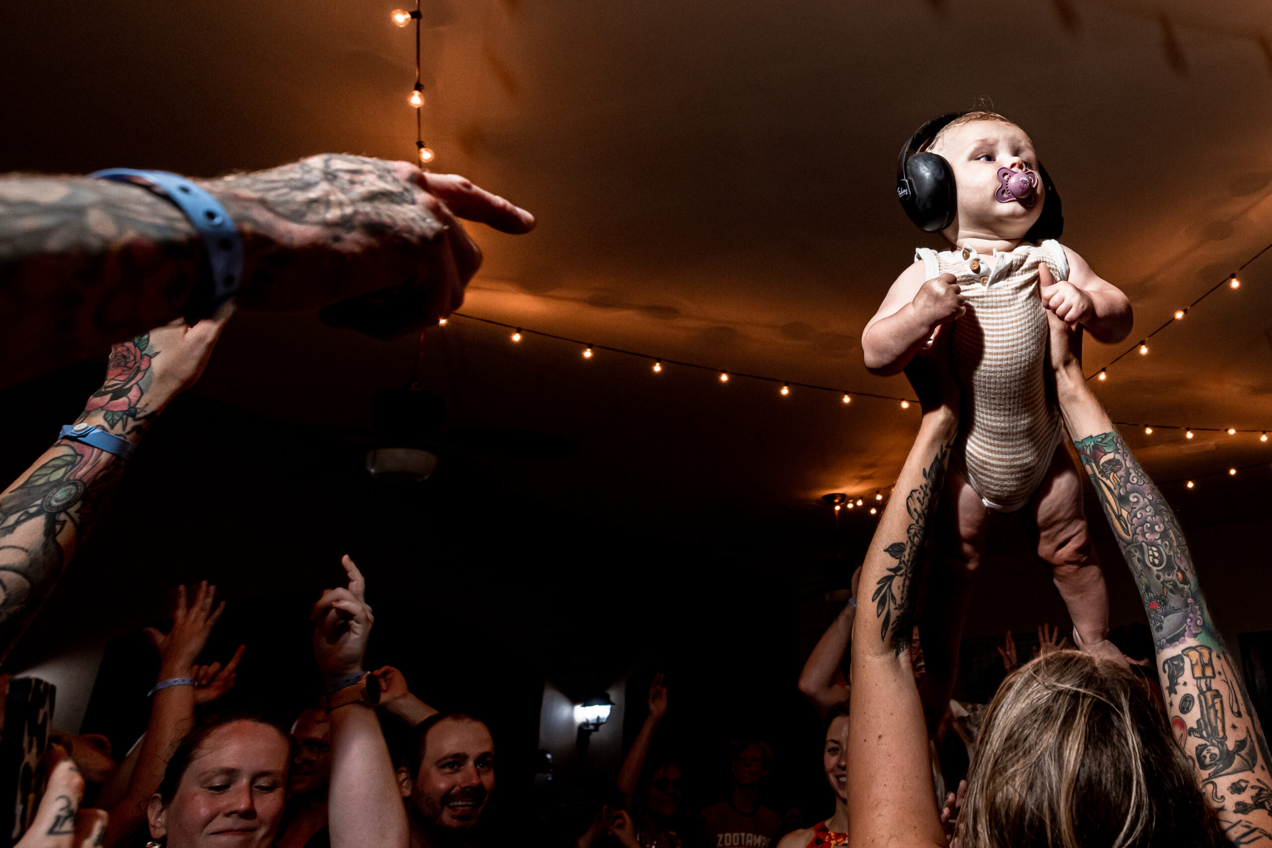 A small baby with earphones is raised above the reception crowd as guests point to her for attention.