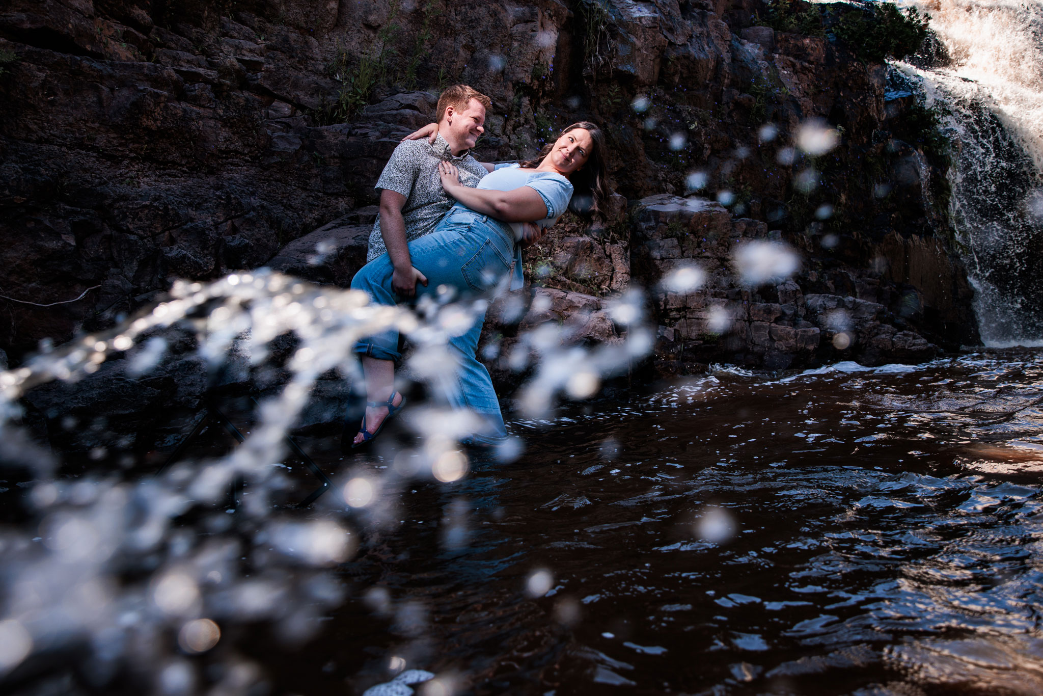 A man dips a woman in the water by a waterfall.