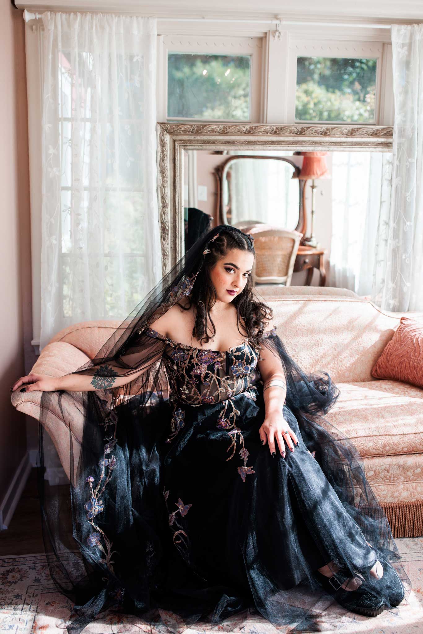 A gothic bride dressed in black ornate wedding dress sits on a pink couch at Camrose Hill Venue.