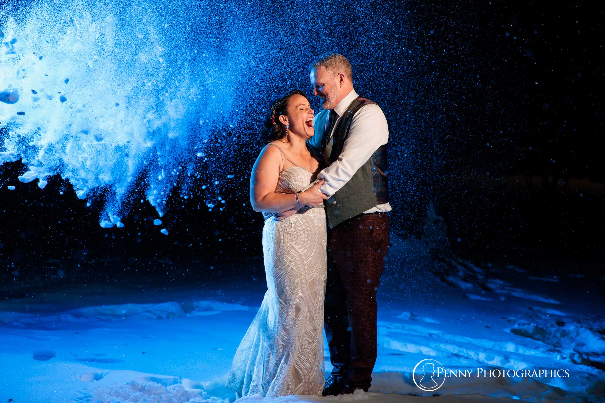 A bride and groom portrait lit up blue with snow falling down on them.
